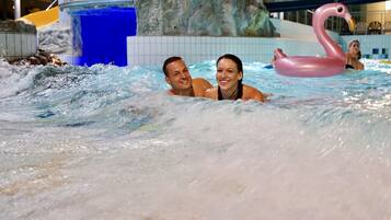 Una pareja en un jacuzzi con un flotador de cisne rosa, rodeada de fuentes de agua y una pared de azulejos.