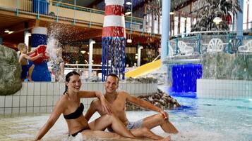 Un couple est assis près d’un élément décoratif aquatique dans un espace piscine intérieure, avec un phare en guise de décoration et un toboggan en arrière-plan.
