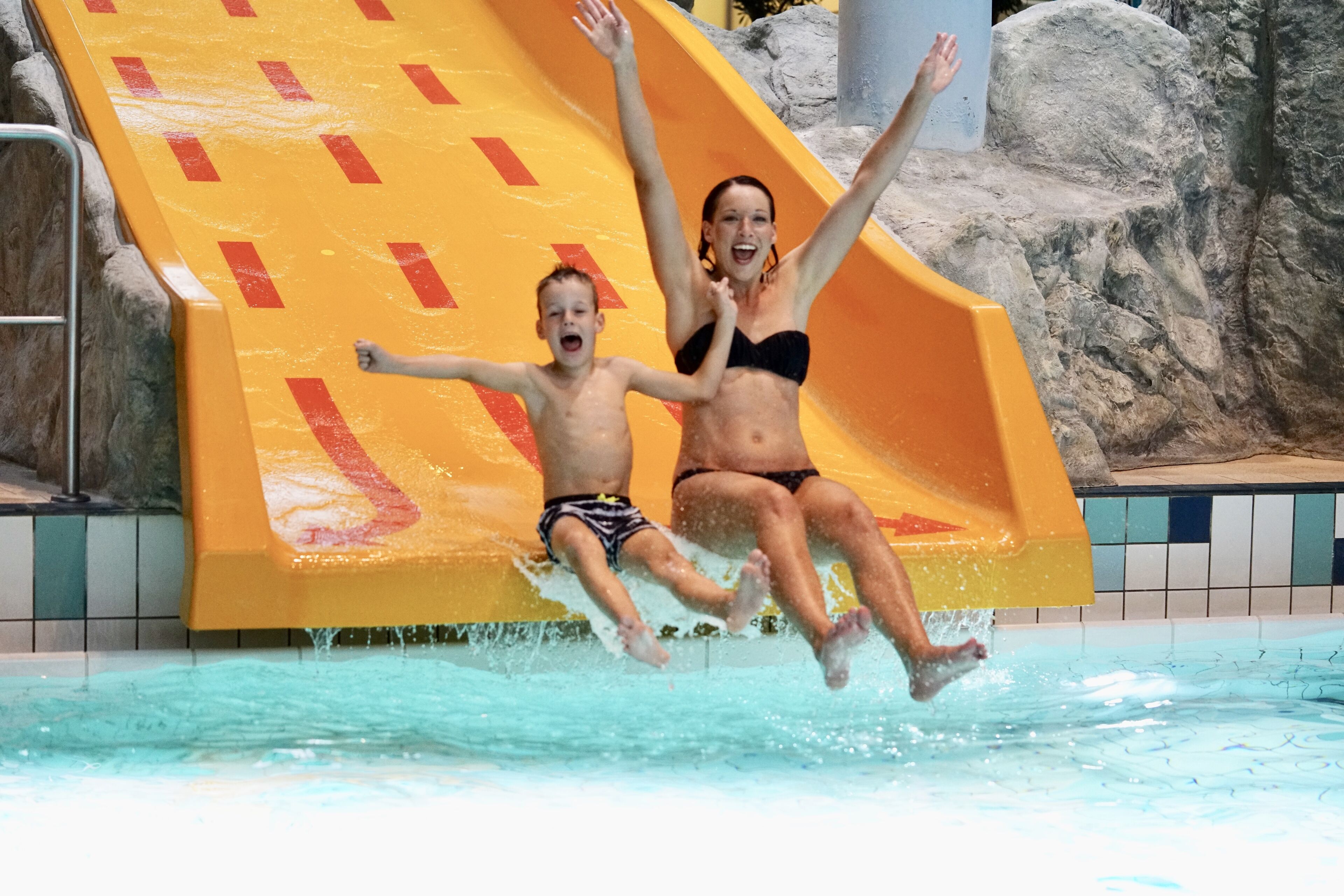 A water slide with a yellow and red design, a pool, and rocky terrain in the background.