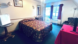 Habitación de hotel con cama, escritorio, silla, televisor, ventana con cortinas y balcón.