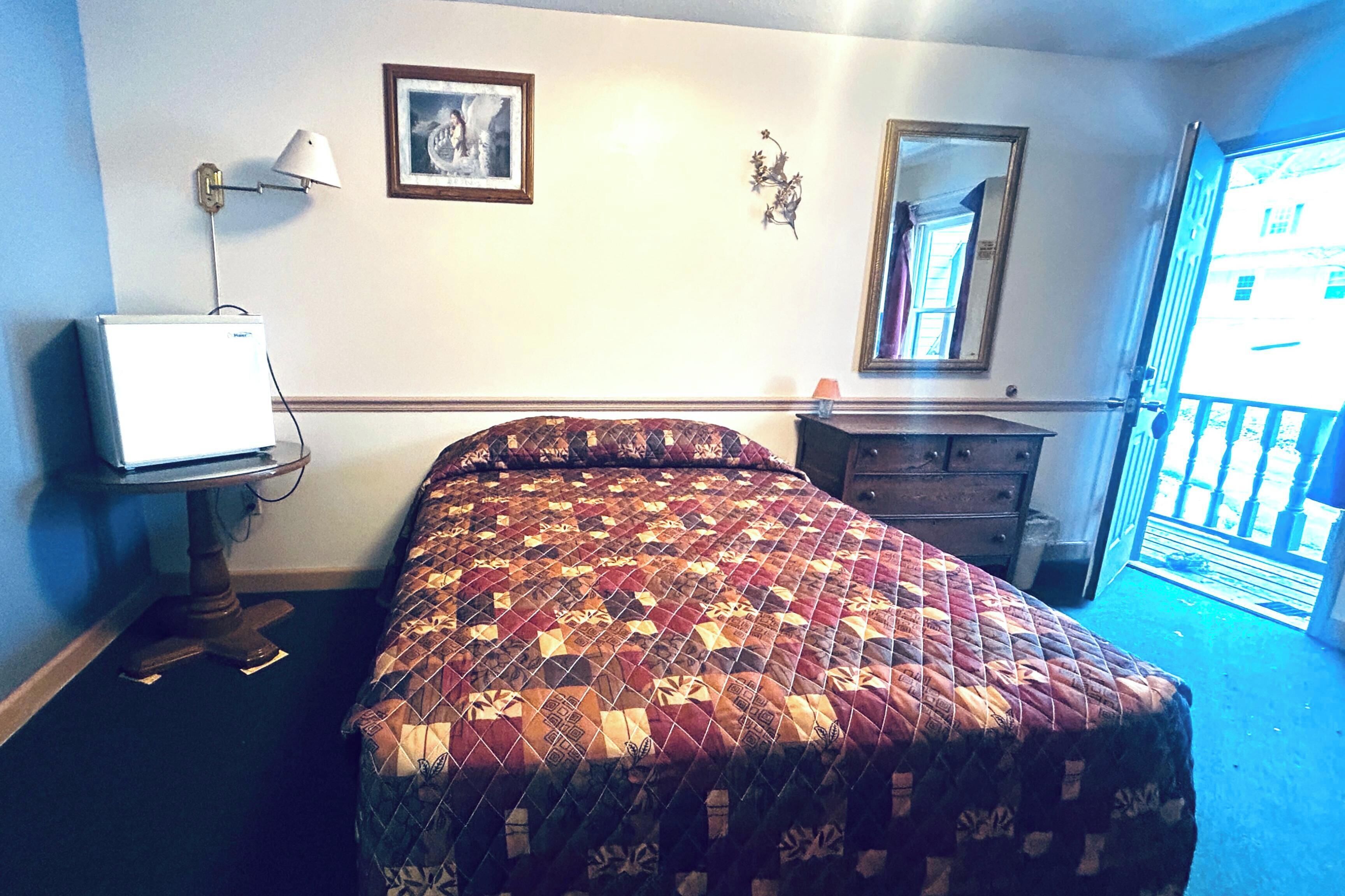 A bedroom with a bed, a desk, a lamp, a dresser, a picture on the wall, and a mirror.