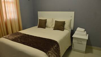 A neatly made bed with a patterned bedspread, two pillows, and a white nightstand with a folded towel.