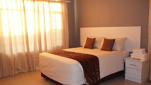 Una cama bien hecha con ropa de cama blanca y marrón, una mesita de noche con toallas dobladas y cortinas translúcidas que dejan entrar luz natural.