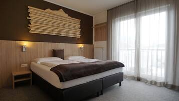 Ein modernes Hotelzimmer mit einem großen Bett, einem Holzkopfteil, einem kleinen Nachttisch und Blick auf die Stadt durch transparente Vorhänge.