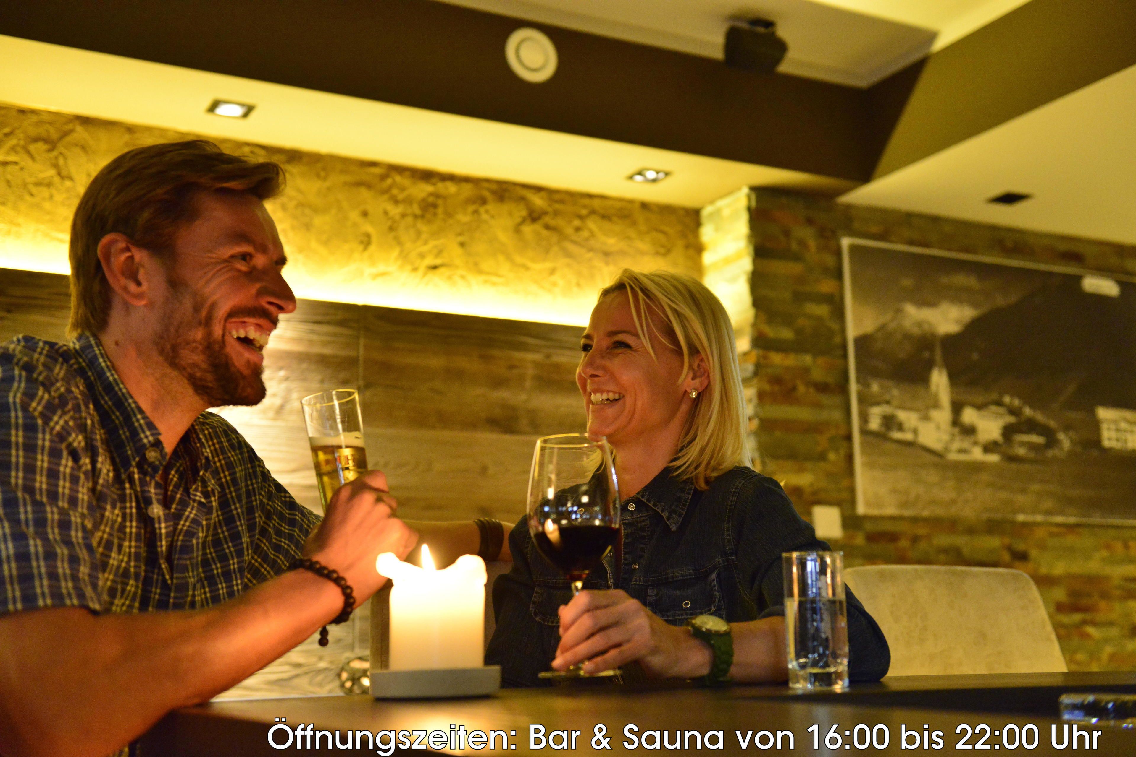 Un homme et une femme trinquent avec des verres de vin à une table où brûle une bougie, dans une pièce dont un mur est en bois et où est accroché un tableau encadré.