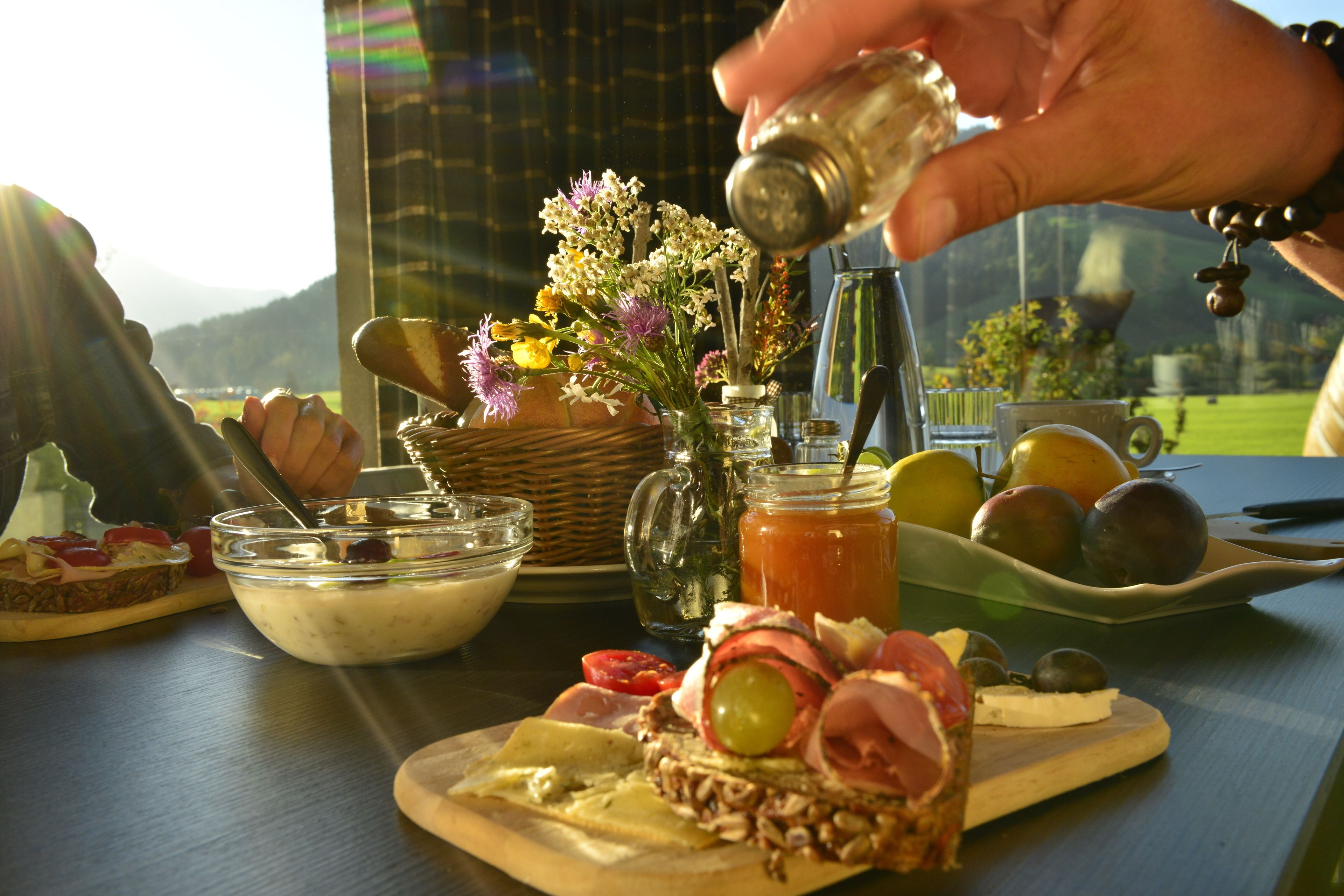 Ein Tisch mit verschiedenen Speisen, darunter ein Sandwich, Käse, Oliven und eine Schüssel Suppe. Dazu steht eine Vase mit Blumen und ein Krug mit Zitronenlimonade.