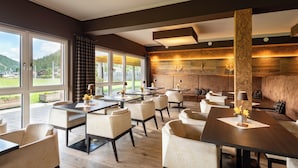 Un restaurante moderno con varias mesas y sillas, ventanas amplias que ofrecen vistas al exterior y una pared revestida de madera que crea un ambiente cálido.