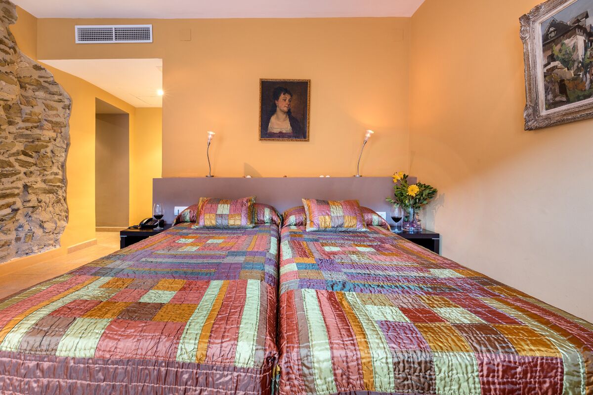 A bedroom with two beds, a stone wall, a painting, and a vase with flowers.
