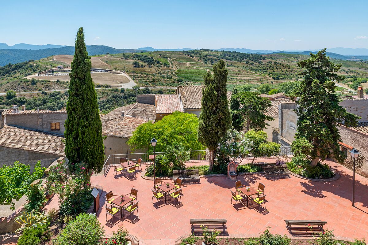Un patio al aire libre con mesas y sillas, rodeado de vegetación y edificios con tejados de teja.