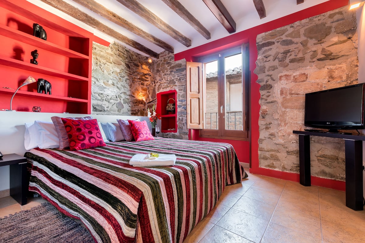 A bedroom with a bed, red shelving, a stone wall, a television, and a balcony door.