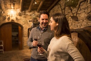 Una pareja sosteniendo copas de vino en una habitación con paredes de piedra, una puerta de madera y un colgante de luz.