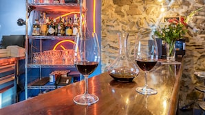 Una zona de bar con mostrador de madera, dos copas de vino, una jarra decantadora y una estantería con botellas y copas.