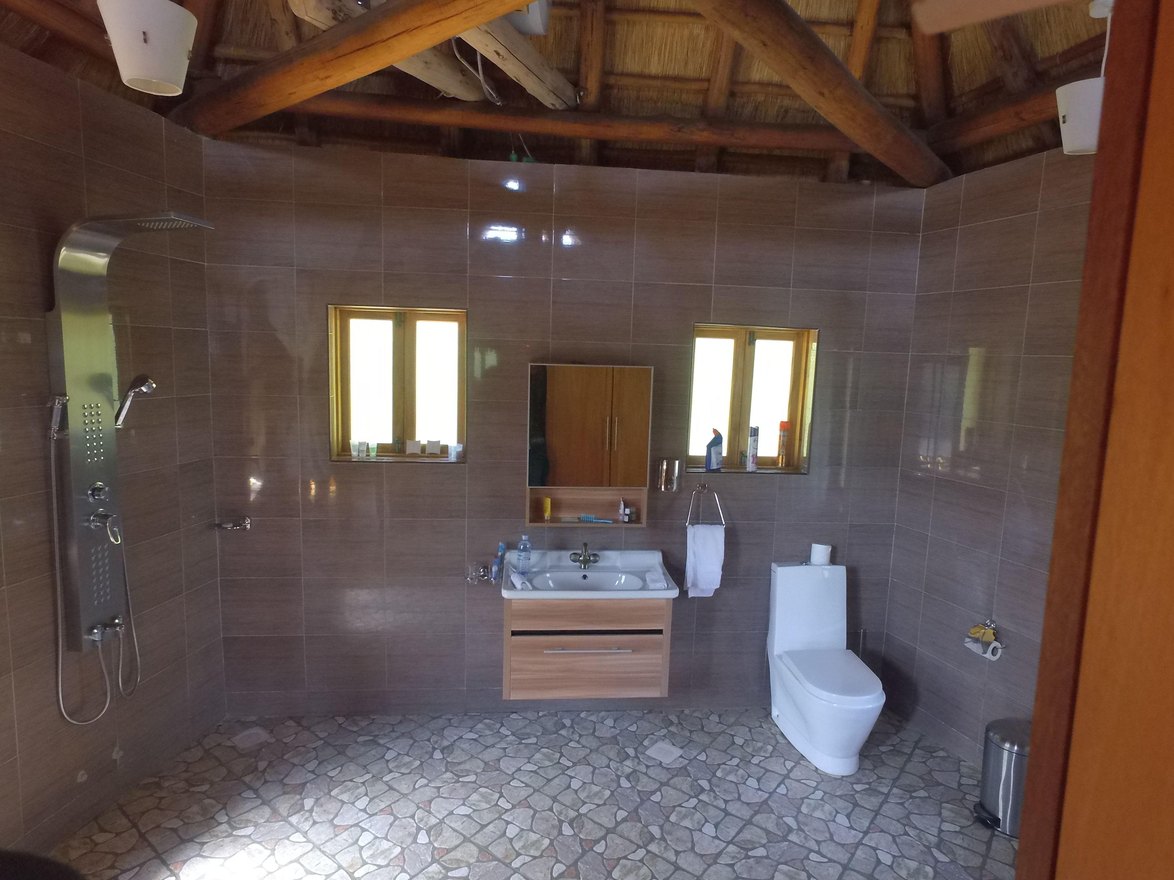 Un baño con ducha, lavabo e inodoro, con suelo de baldosas con diseño de piedra, vigas de madera en el techo y dos espejos adosados a la pared.