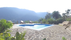 A swimming pool with lounge chairs, surrounded by stone flooring and greenery, with mountains in the background.