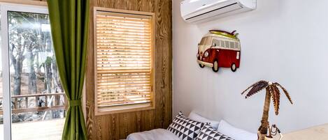 Une chambre à coucher avec un lit, une fenêtre avec des stores, un climatiseur fixé au mur, et une décoration murale représentant une voiture et un palmier.