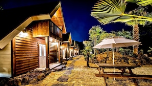 A row of wooden cabins with a stone pathway leading to a table and umbrella. - Browncabin Pension (Jeju City)