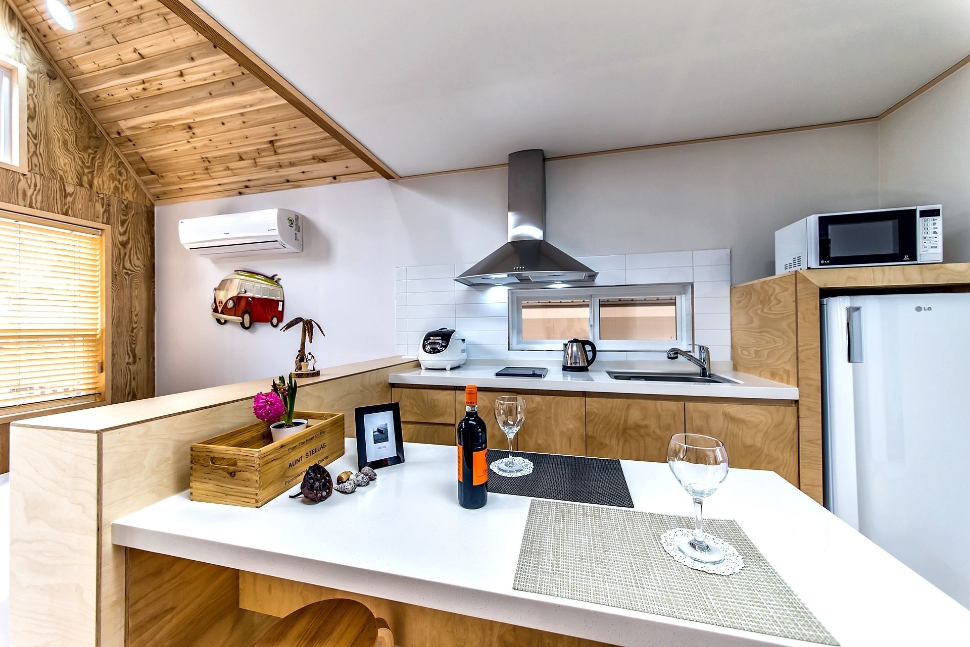 A compact kitchen with a microwave, stove, and refrigerator. A wooden counter with a picture frame, bottle, and glass is in the center.