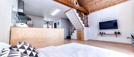 A modern bedroom with a wooden ceiling, a bed with patterned pillows, a TV on the wall, and a staircase leading to an upper level.