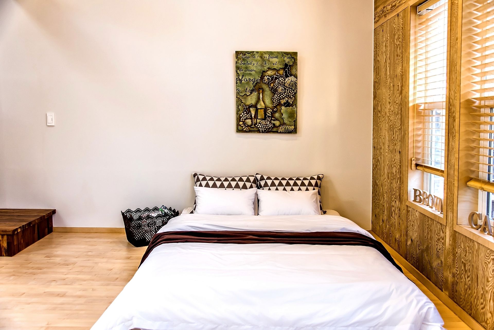Un dormitorio con piso de madera, una cama con ropa blanca, un cojín a cuadros en blanco y negro, un cabecero de madera y una ventana con persianas.