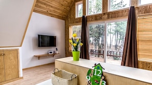 A room with a wooden ceiling, a TV mounted on the wall, a window with curtains, a shelf with a plant, a clock, and a woven basket. - Browncabin Pension (Jeju City)