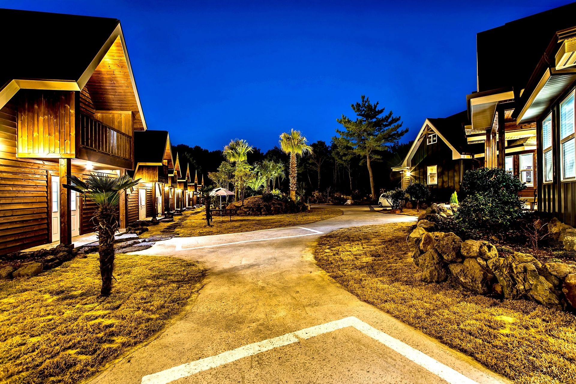 A well-lit, tree-lined driveway leading to a row of wooden houses with a clear path and no visible vehicles.