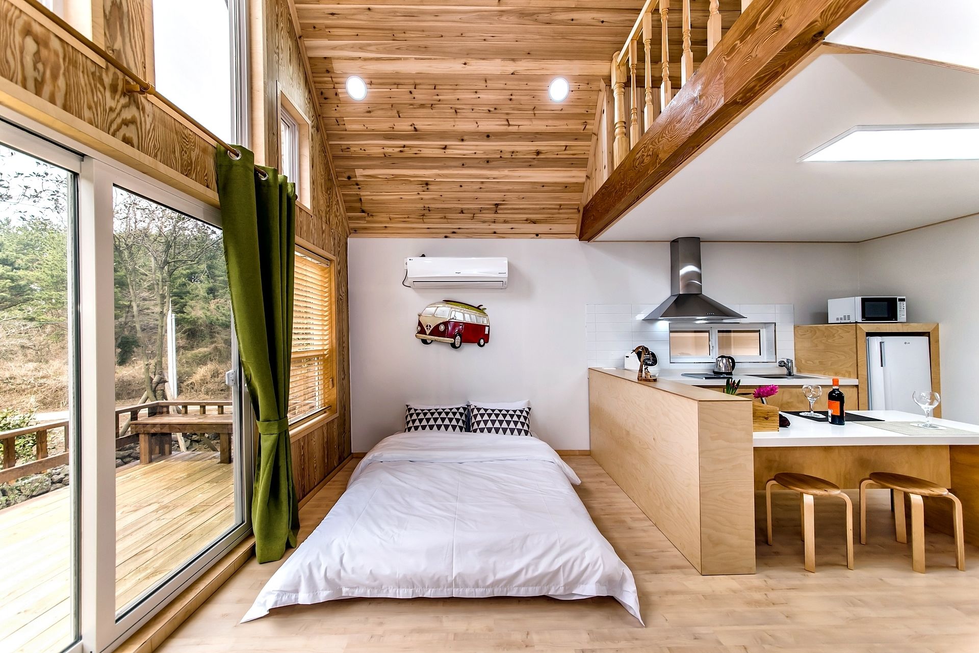 A bedroom with a large window, a bed, a small kitchenette, and a wooden deck outside.