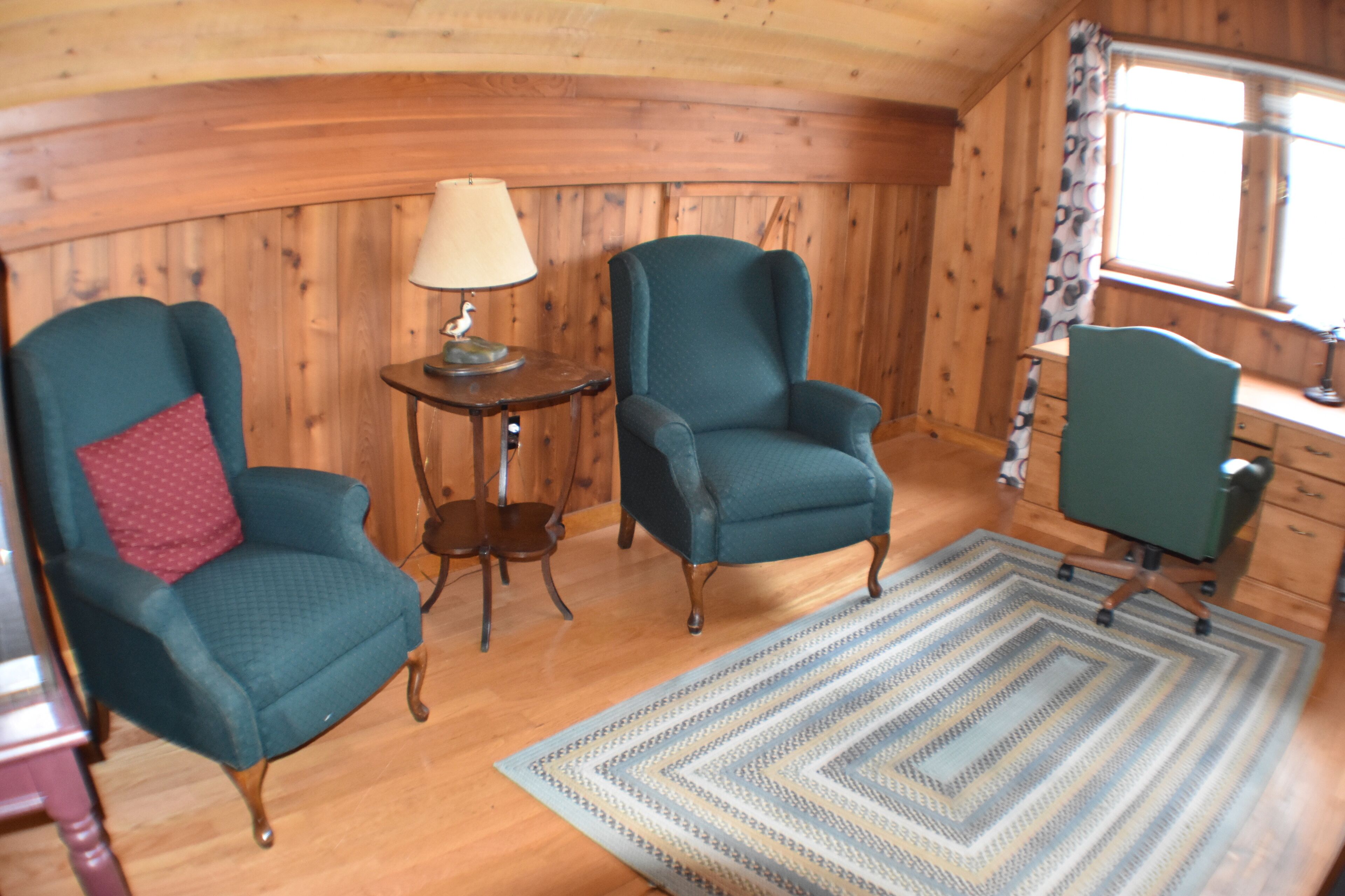 A room with wooden walls and a wooden floor, featuring two teal armchairs, a small table with a lamp, and a patterned rug.