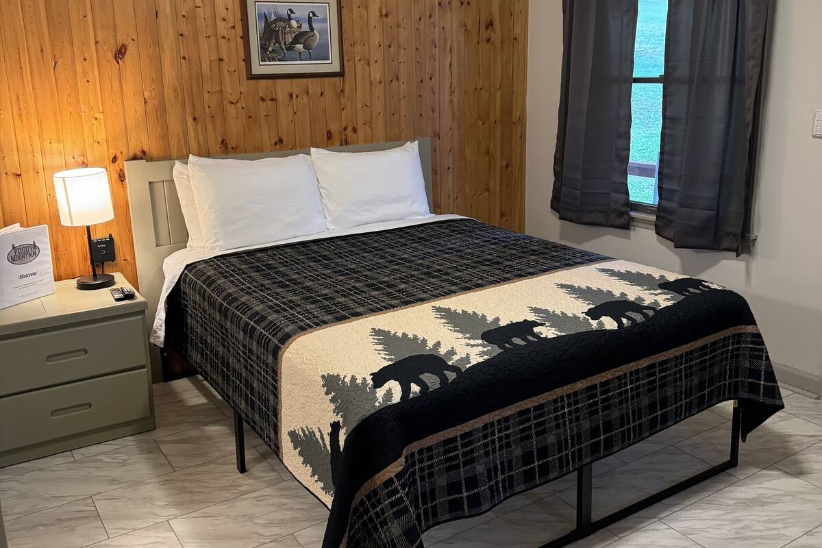 Une chambre avec un mur lambrissé en bois, un lit recouvert d’une couvre-lit à motifs, une table de chevet avec une lampe et une fenêtre avec des rideaux.