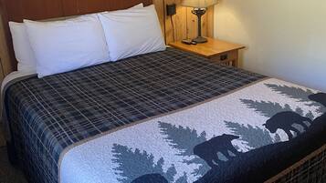 A bed with a patterned bedspread, a wooden headboard, a nightstand with a lamp, and a framed picture on the wall.