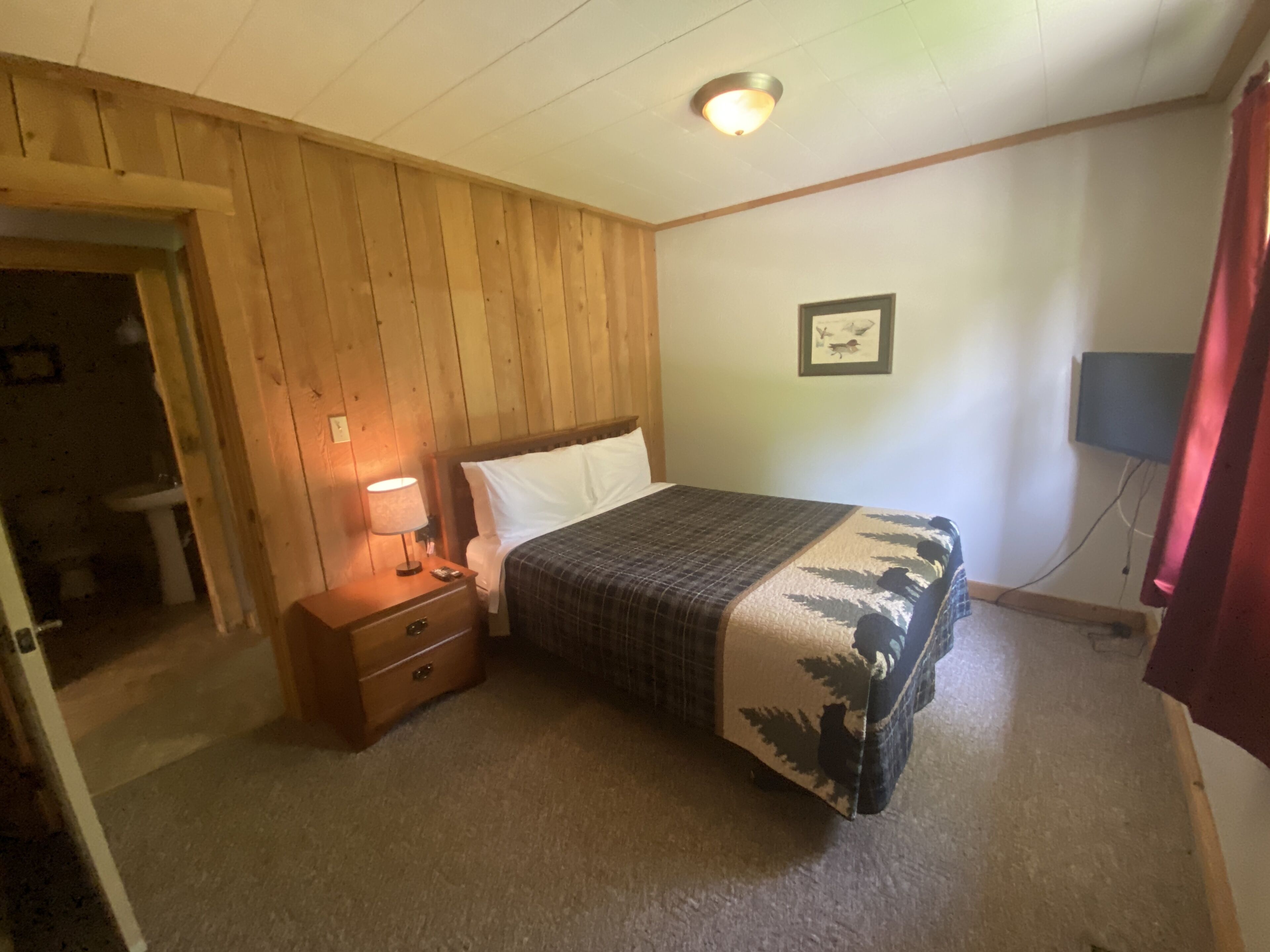 A bedroom with a wooden headboard, a bed with a patterned bedspread, a nightstand with a lamp, and a wall-mounted TV.