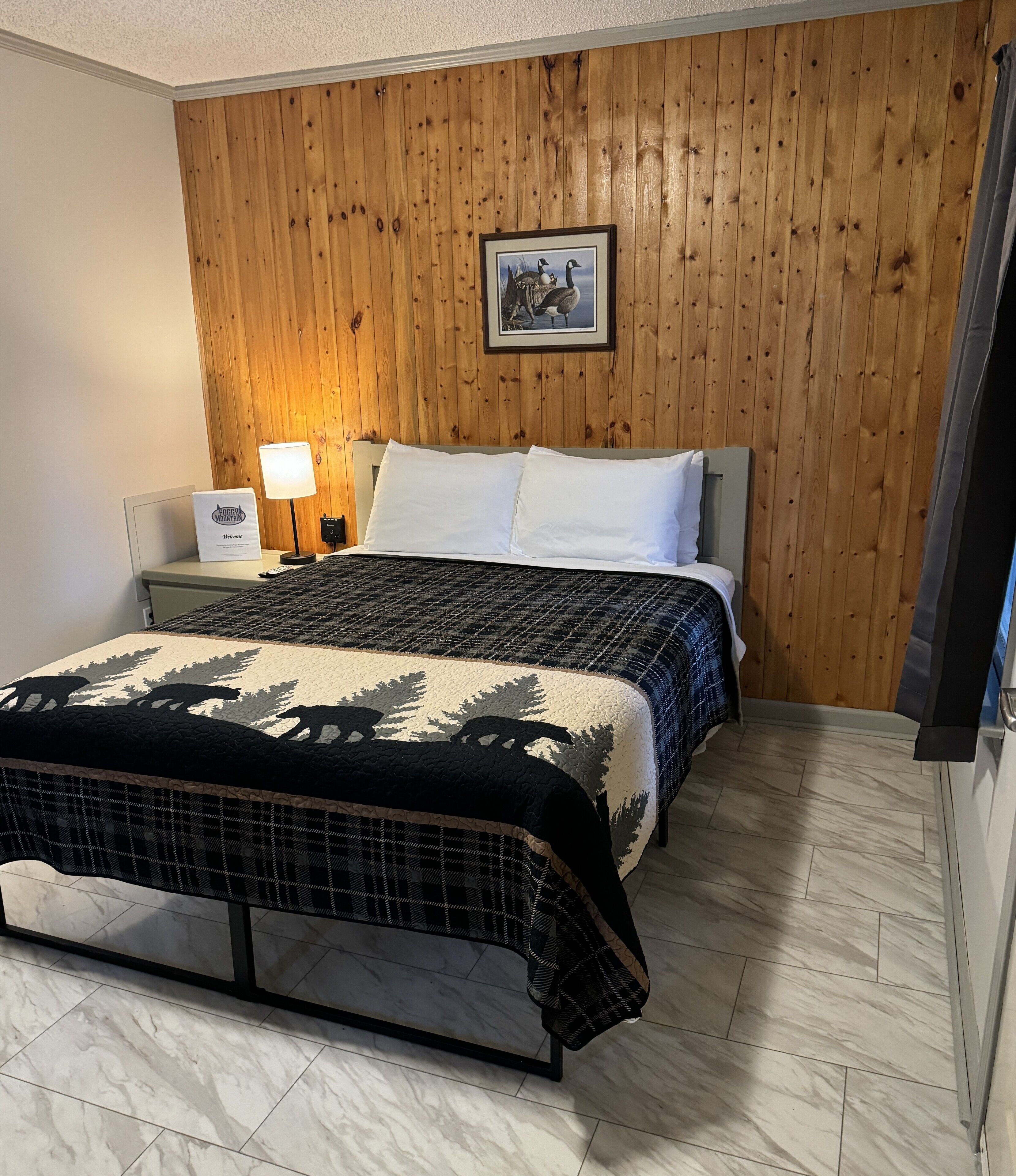 Ein Hotelzimmer mit einer holzgetäfelten Wand, einem Bett mit gemustertem Überwurf, einem Nachttisch mit Lampe und einem gerahmten Bild an der Wand.