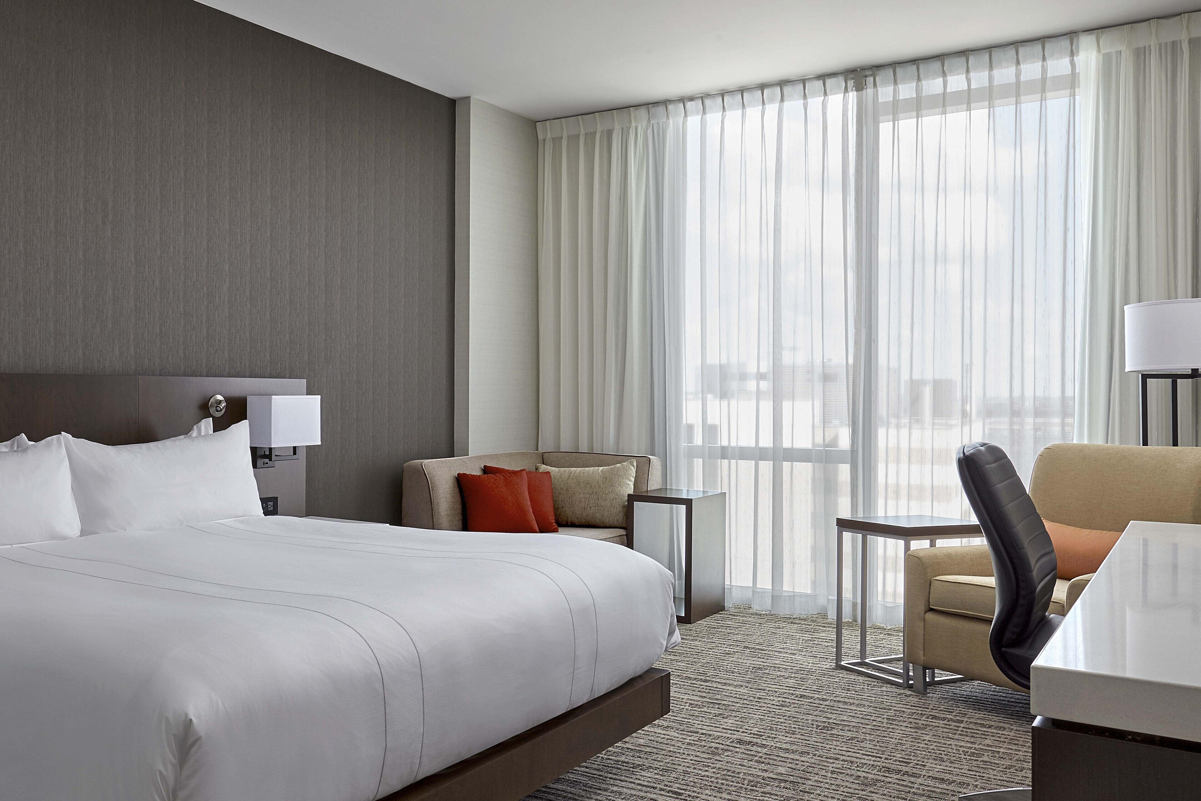 A hotel room with a large bed, a sofa, a chair, a small table, and a lamp.