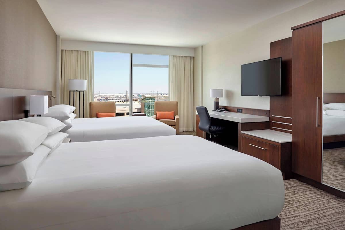 A hotel room with two beds, a desk, a chair, a TV, and a large window with a city view.