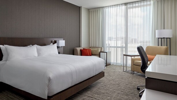 A hotel room with a large bed, a desk, a chair, and a view of the city through sheer curtains. - Calgary Airport Marriott In-Terminal Hotel (Calgary)