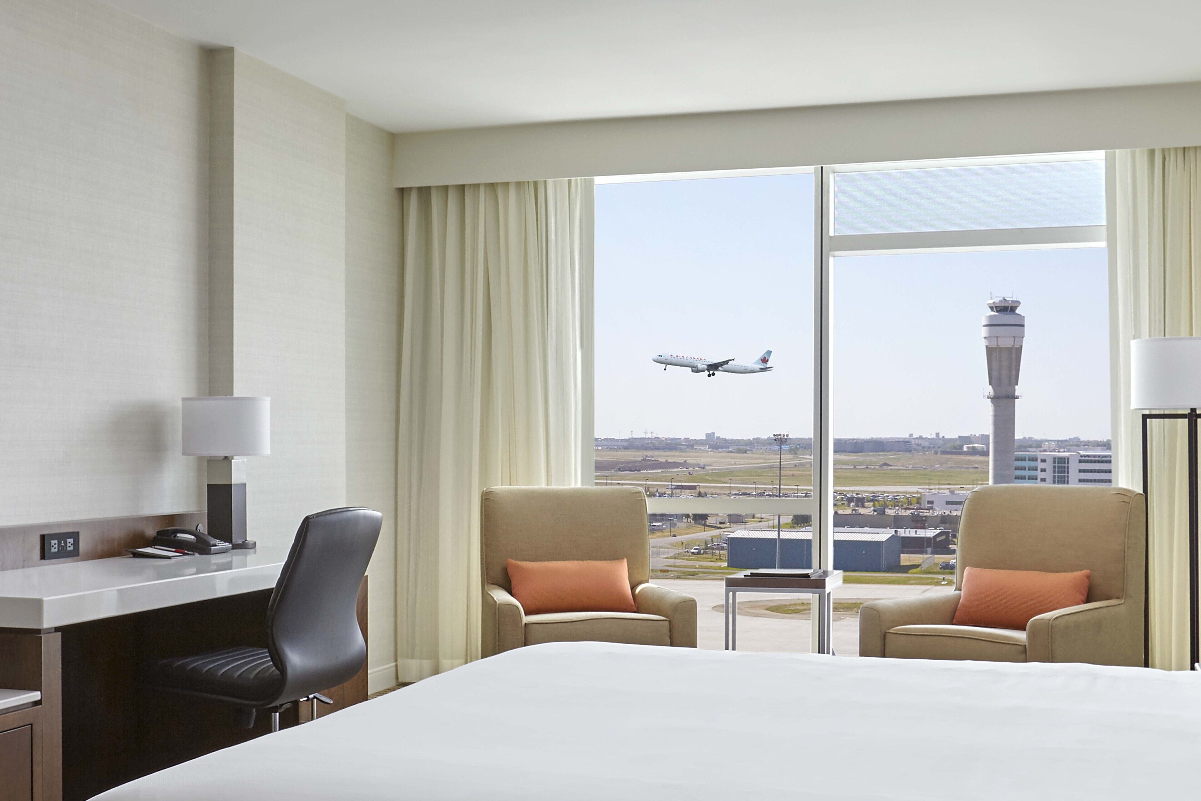 A hotel room with a large bed, two armchairs, a desk with a lamp, and a view of an airport with an airplane and a control tower.