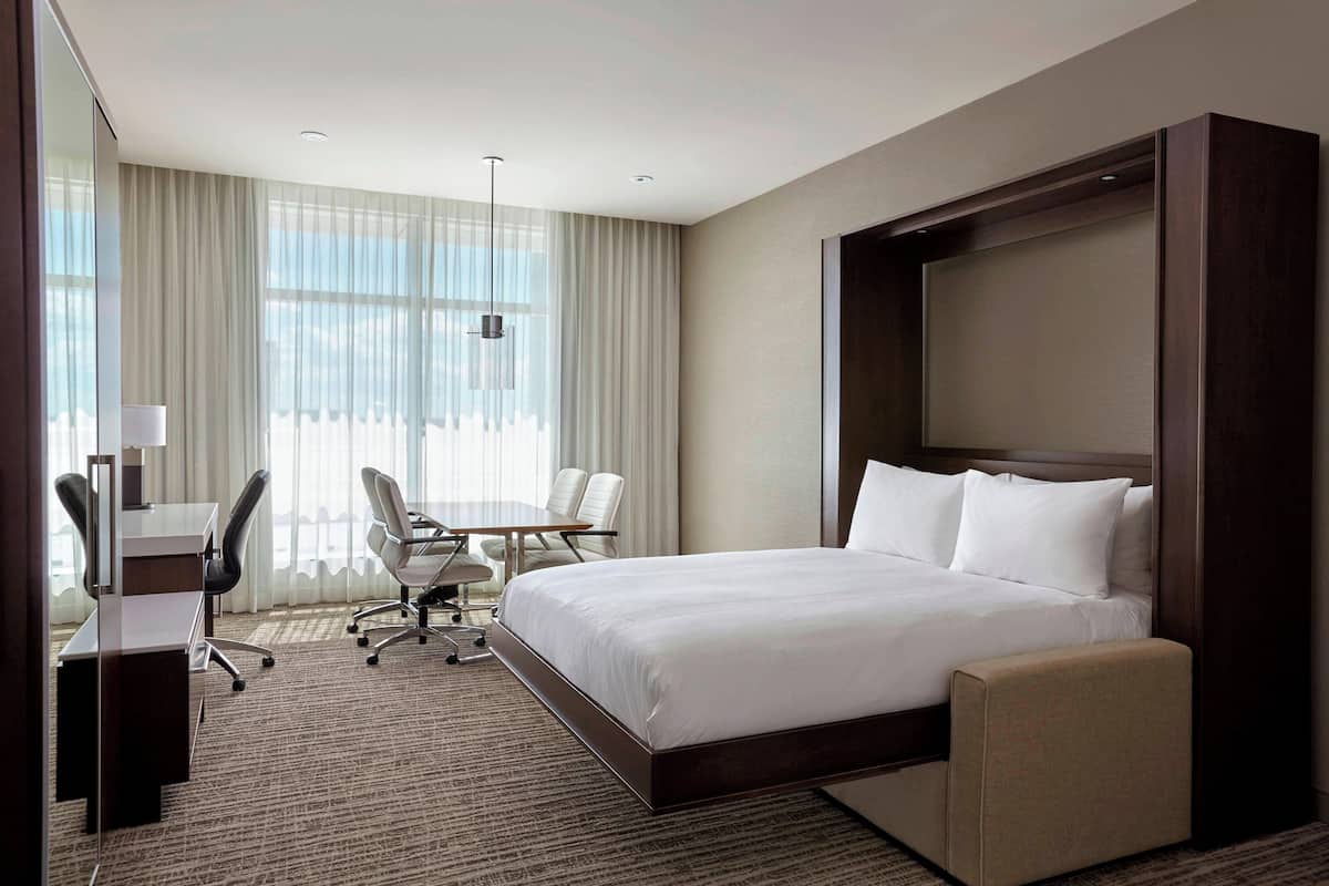 A hotel room with a large bed, a desk with a chair, a small table, and a large window with curtains.