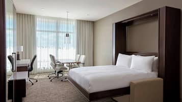 Habitación de hotel con una cama grande, un escritorio con silla, una mesita pequeña y un ventanal con cortinas.