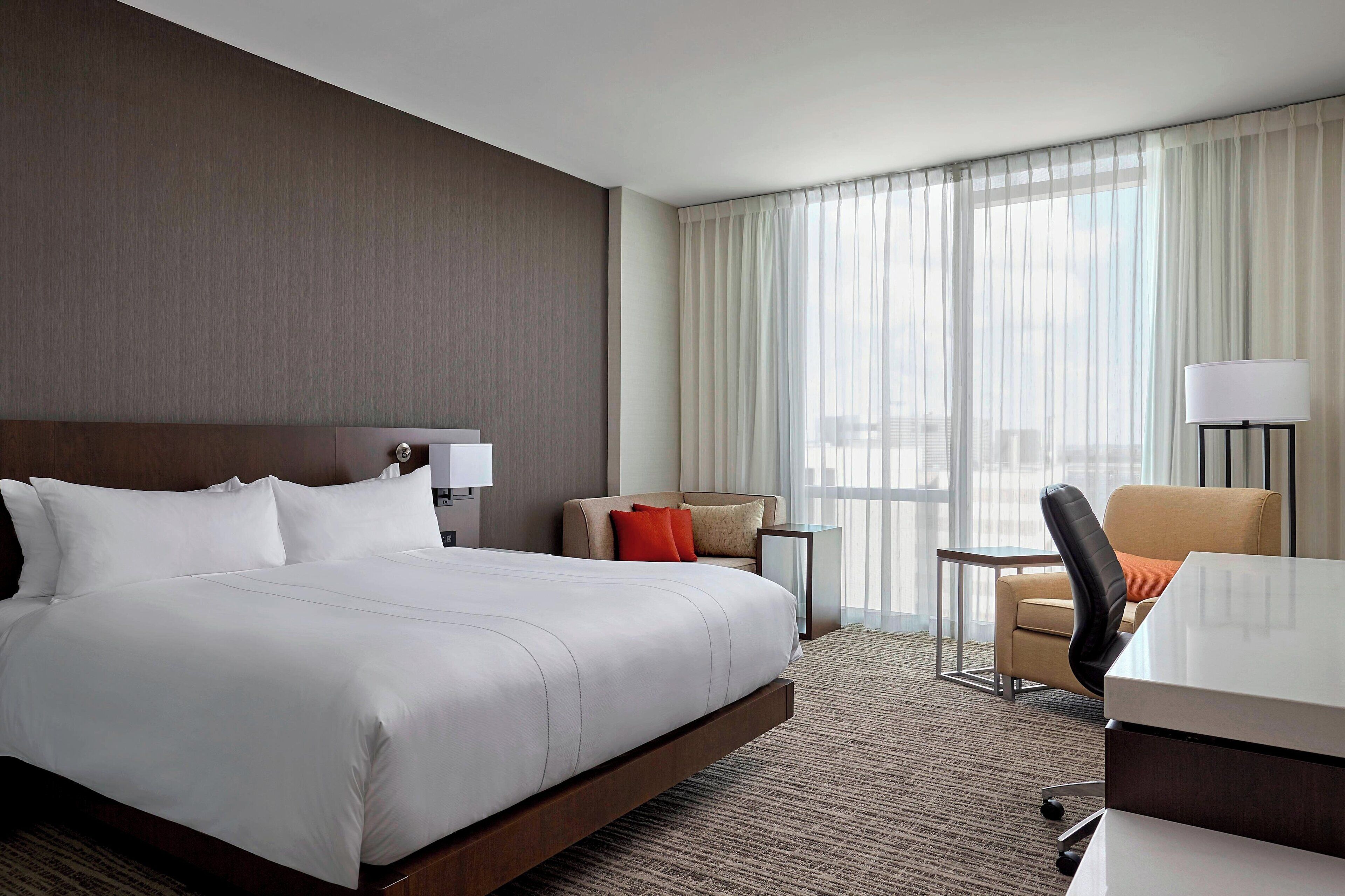 Habitación de hotel con una cama grande, un escritorio, una silla y vistas de la ciudad a través de cortinas translúcidas.