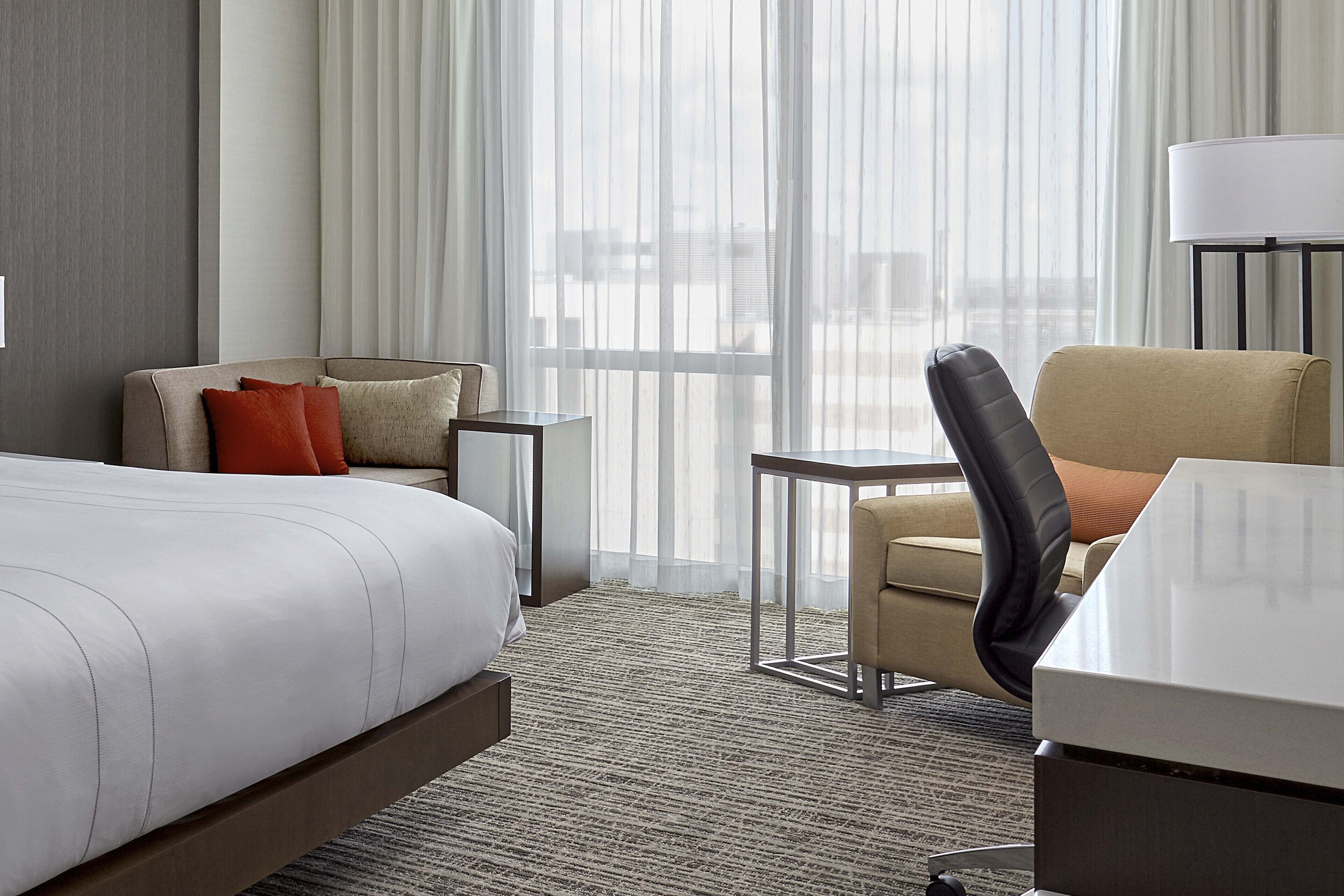 A hotel room with a bed, a desk, a chair, a small table, and a view of the city through large windows.