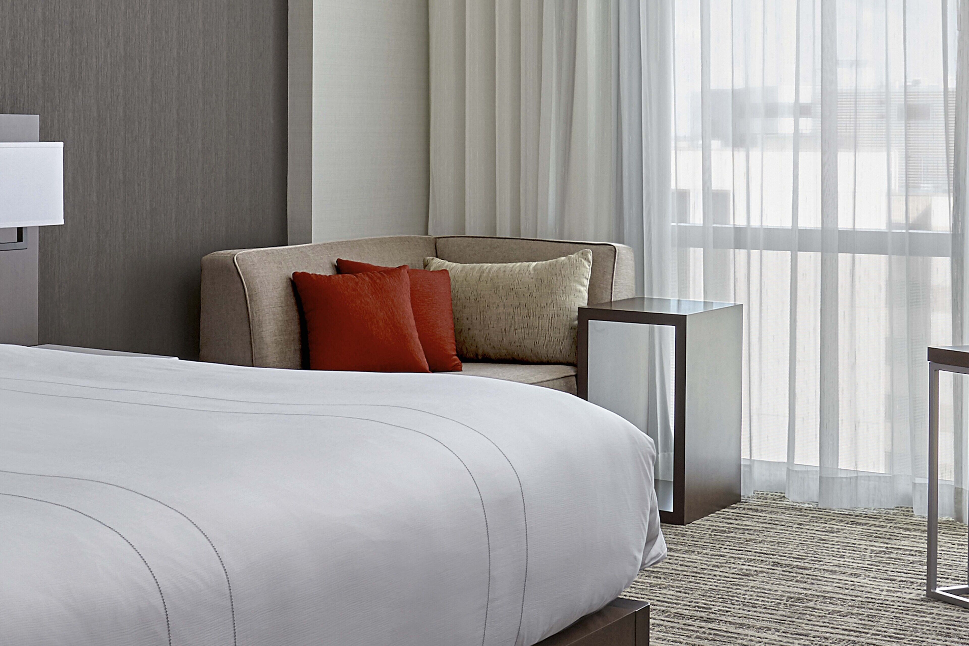 A hotel room with a large bed, a sofa with cushions, a small table, and sheer curtains.