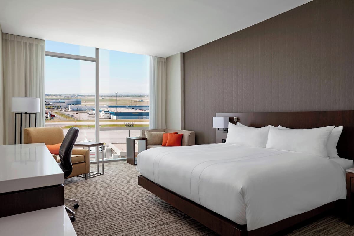 A hotel room with a large bed, a desk, a chair, and a view of an airport.