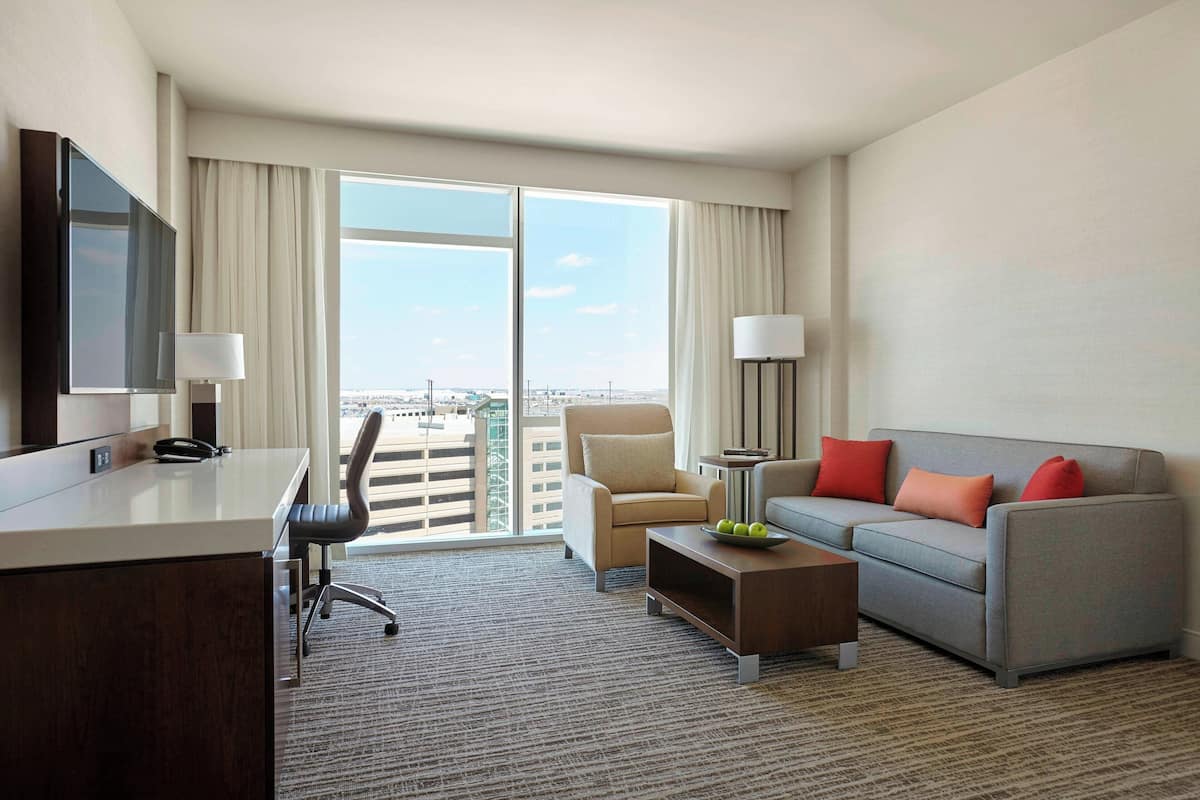 A hotel room with a desk, chair, sofa, and a large window offering a city view.