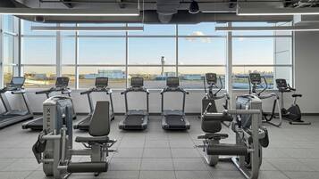 Ein Fitnessstudio mit Laufbändern, Crosstrainern und Gewichten, mit Blick auf einen Flughafen und Stadtpanorama.