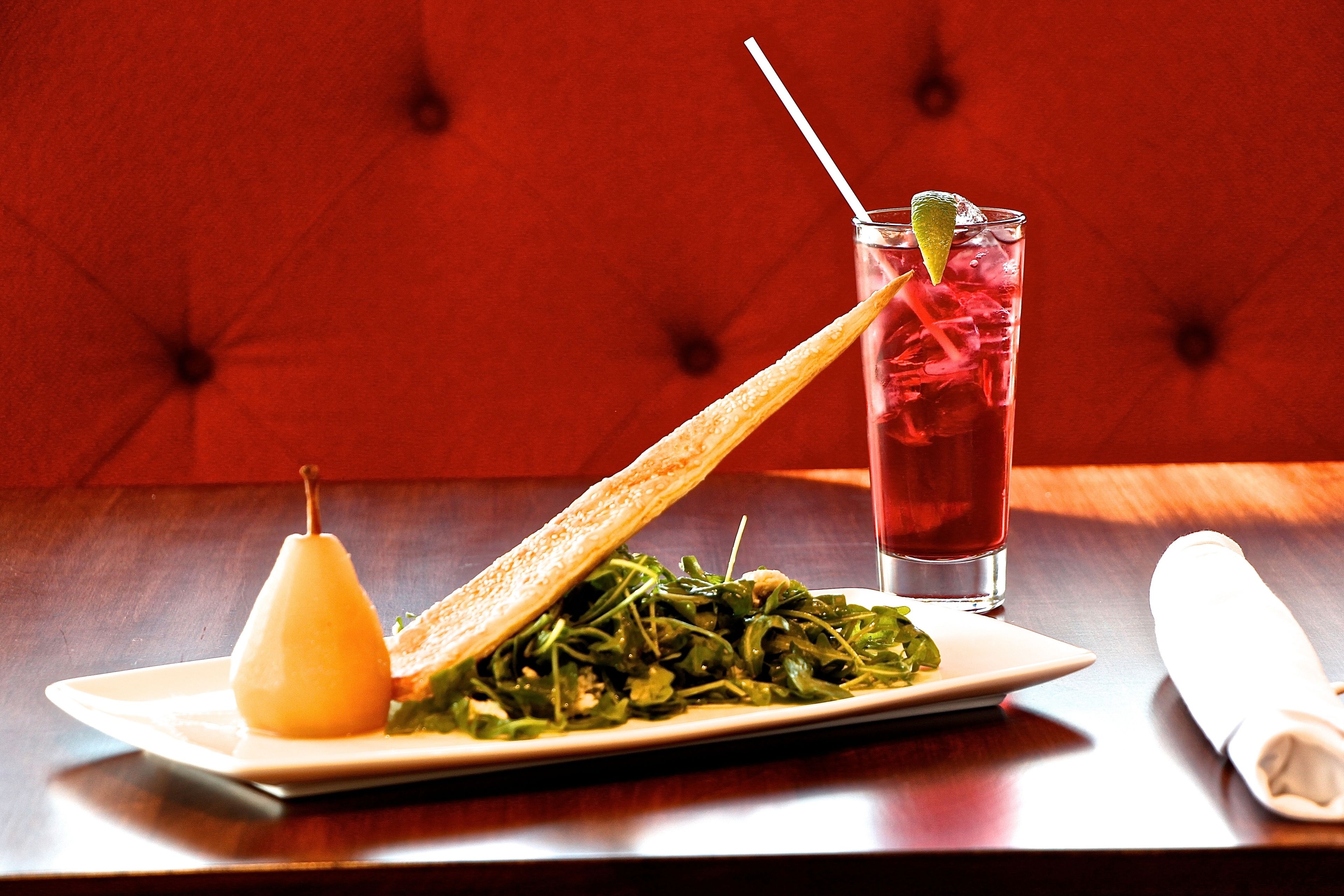 A plate of greens with a pear and a slice of bread, a glass of red drink with a straw and lime, and a folded napkin on a wooden table.