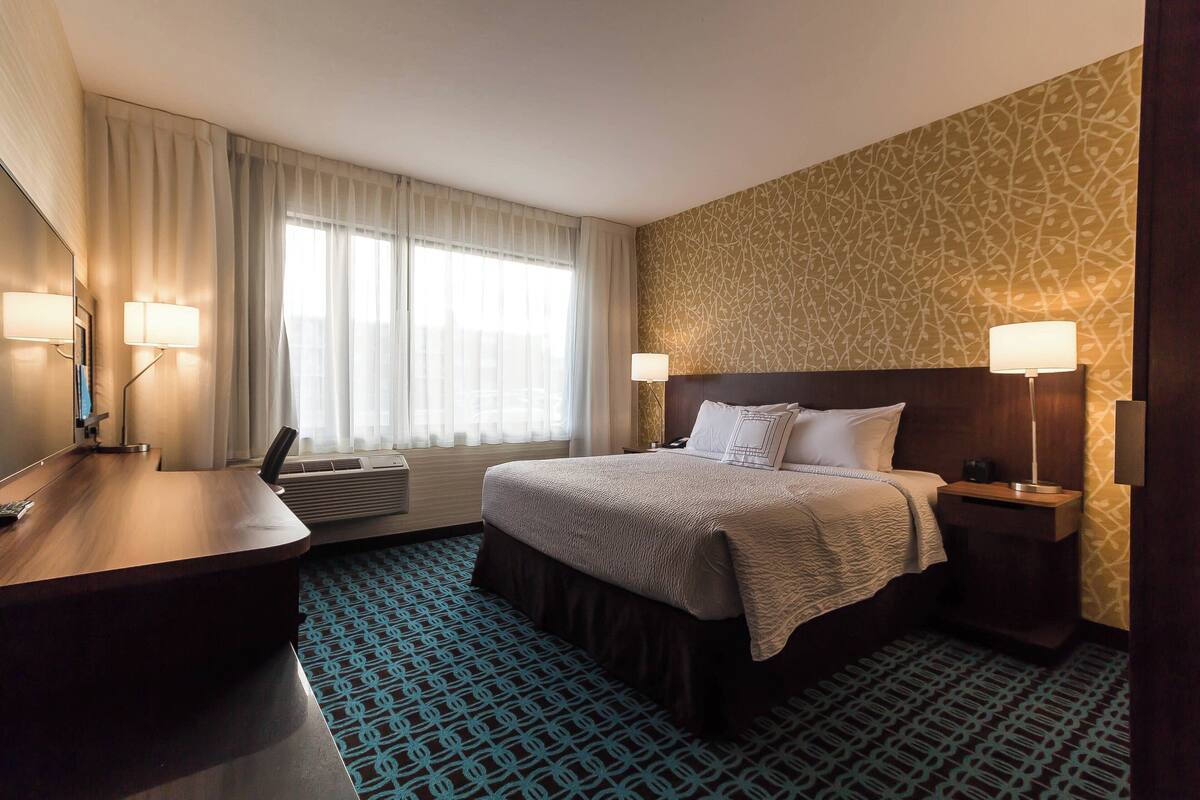 A hotel room with a bed, bedside tables, a desk, and a large window with curtains.