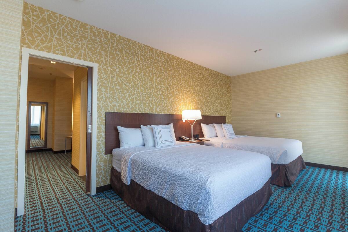A hotel room with two beds, a nightstand with a lamp, a patterned carpet, and a door leading to another room.