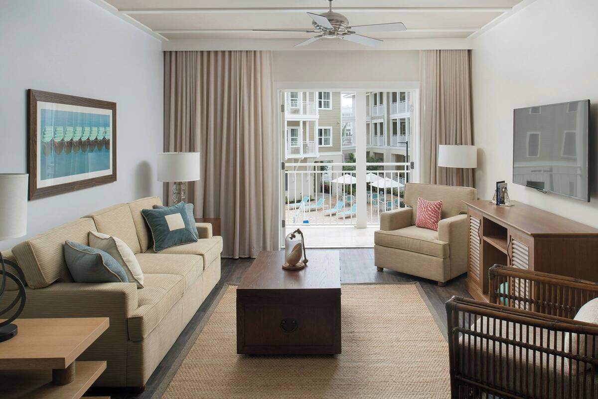 Een hotelkamer met een bank, fauteuil, houten salontafel en een wieg. Er is een televisie aan de muur en een balkon met buitenmeubilair.