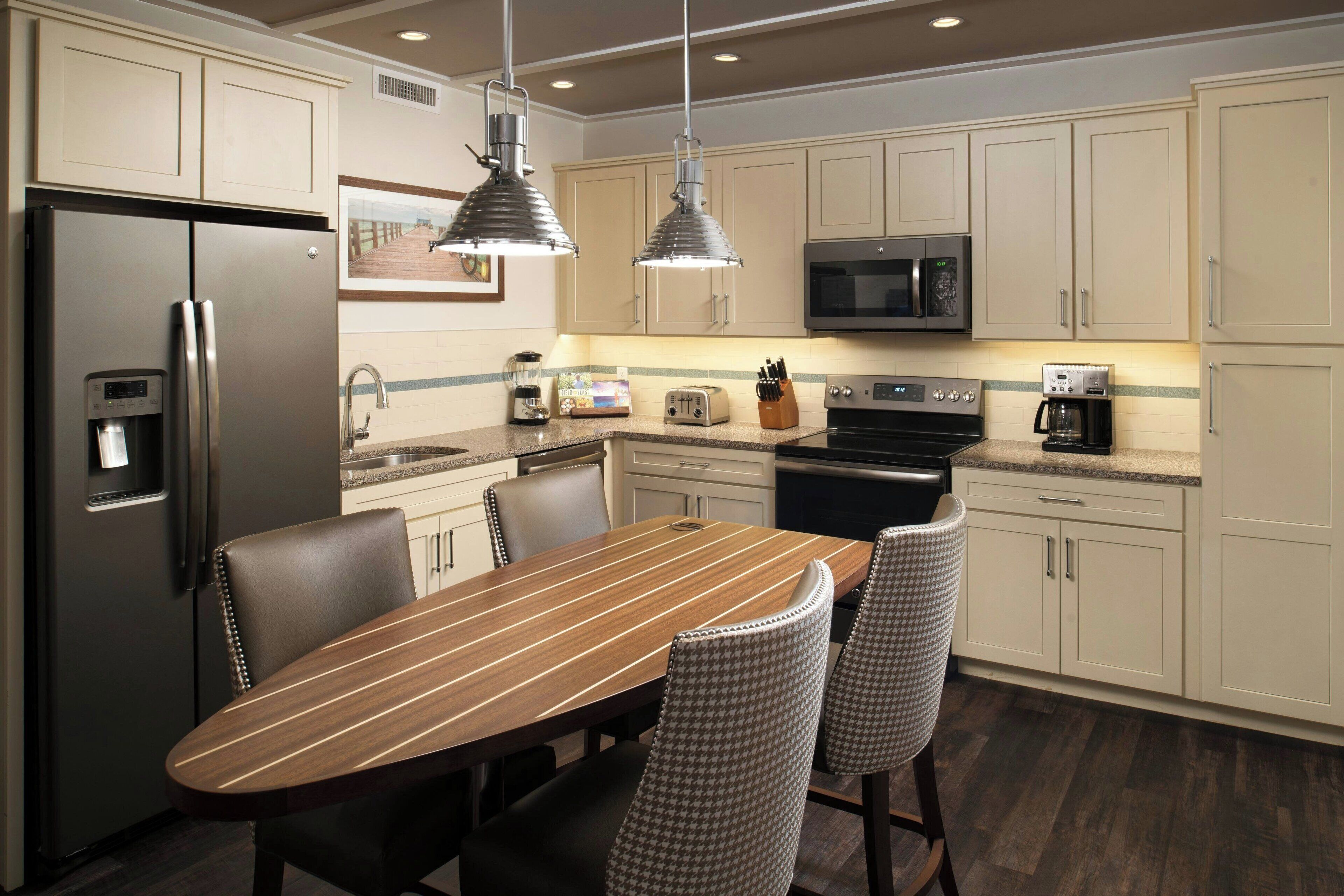 A modern kitchen with a stainless steel refrigerator, a microwave, a coffee maker, and a toaster. There is a wooden dining table with four chairs.