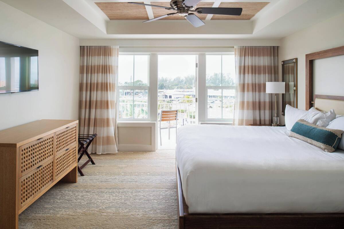 Een hotelkamer met een bed, een houten dressoir, een plafondventilator en een groot raam met gordijnen.
