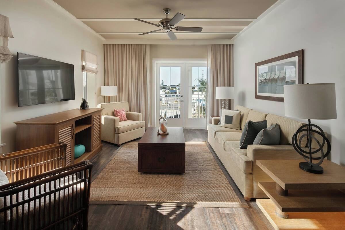 Hotelkamer met flatscreen televisie, plafondventilator, groot raam met gordijnen, een bank, salontafel en een houten kast.